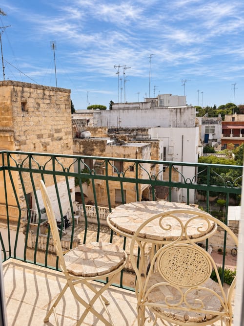 Balcone con sedie color crema e tavolo con vista sui tetti storici di Lecce, edifici in pietra antica e cielo azzurro