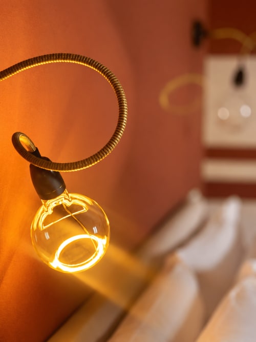 Warm glowing pendant lamp with brass flexible arm in HABITARE Lecce room, creating ambient lighting against orange wall