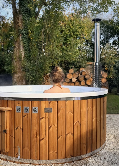 Hôte se relaxant dans un bain à remous en bois à la Maison Mansel en Mesnil-en-Ouche, entouré d'arbres d'automne et de bois de chauffage