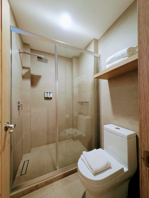 Baño moderno en el hotel Casa Donceles de la Ciudad de México con ducha de vidrio, inodoro blanco, estantería de madera con toallas y tonos beige