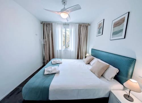Modern bedroom in Tenerife apartment with teal headboard, white bedding, ceiling fan, and curtained window