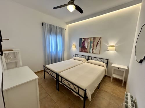 Dormitorio moderno en apartamento Judería3, Sevilla, con cama de metal negro, ropa de cama blanca, ventilador de techo, apliques de pared y obras de arte