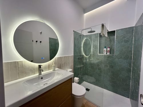Baño moderno en apartamento Judería3, Sevilla, con espejo iluminado redondo, azulejos hexagonales verdes, lavabo blanco y ducha.