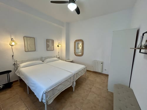 Dormitorio luminoso con cama de estructura metálica blanca, ventilador de techo, apliques de pared y espejos en el apartamento CozyHomeSevilla, Sevilla.