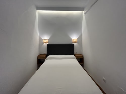 Dormitorio moderno en CozyHomeSevilla, Sevilla con paredes blancas, cabecero negro, mesillas de madera y apliques de luz ambiental