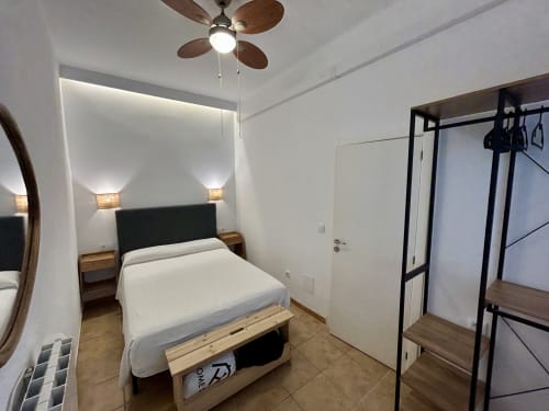 Dormitorio moderno en CozyHomeSevilla, Sevilla con paredes blancas, cama, ventilador de techo, mesitas y armario de metal negro