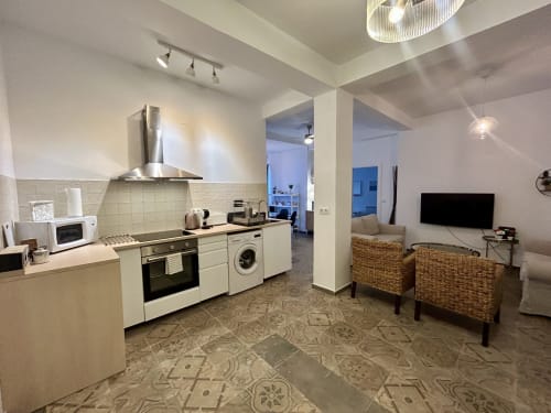 Apartamento moderno de planta abierta en Sevilla con cocina equipada, zona de estar con sillas de ratán, TV y suelo de azulejos con patrón.