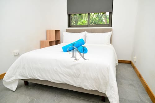 Chambre lumineuse de la villa Nora à Grand Baie avec literie blanche, oreillers bleus, rangements en bois et vue sur le jardin verdoyant