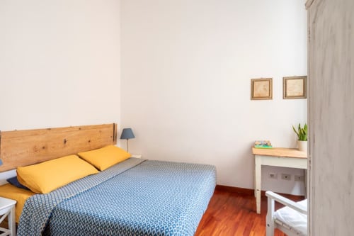Chambre confortable à Rome House Rental avec lit en bois, oreillers jaunes, literie bleue et bureau en bois dans l'appartement Secret Corner