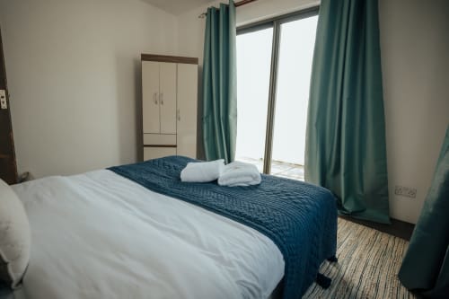 Modern villa bedroom with white and navy bedding, teal curtains, wardrobe, and sliding glass doors opening to pool view at