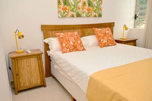 Chambre lumineuse avec lit en bois, oreillers à motifs orangés, draps blancs, lampe jaune et œuvres florales chez Gerards & Angel