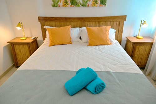 Chambre d'appartement en bord de mer avec lit en bois, coussins jaune or, serviettes turquoise roulées et lampes de chevet