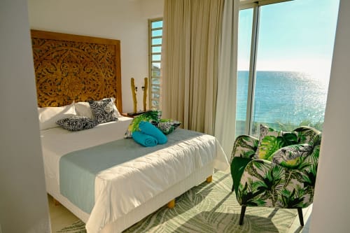 Chambre en bord de mer chez Gerards & Angel avec tête de lit sculptée, vue océan, fauteuil tropical et baies vitrées face à la mer