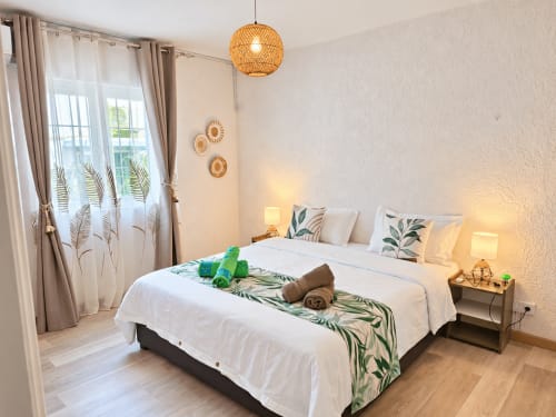 Bright bedroom at Gerards & Angel with white bed, tropical green runner, woven pendant light, and fern-patterned curtains
