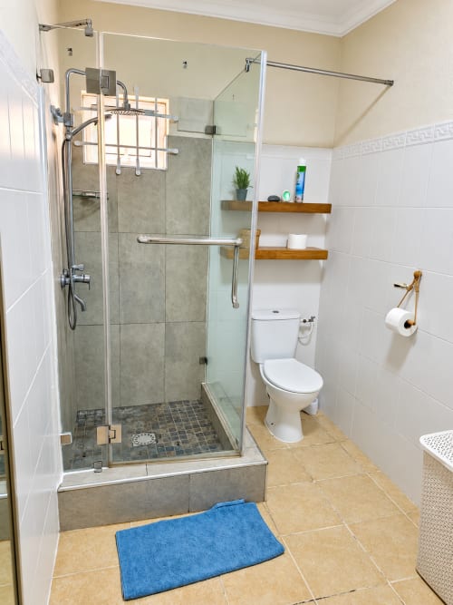 Modern bathroom at Gerards & Angel with glass shower enclosure, white tile walls, wooden shelves, toilet, and blue bath mat