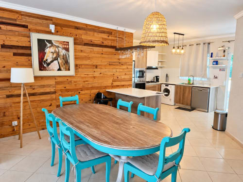 Open-plan kitchen and dining area with turquoise chairs, wooden table, wood-paneled walls, and woven pendant light at Gerards