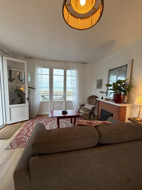 Salon côtier lumineux avec canapé gris, mobilier en bois et vue océan depuis les portes de balcon à Mon Week End A