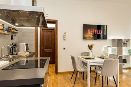 Cucina e zona pranzo di moderno appartamento romano con Netflix sulla TV, tavolo bianco, sedie beige e pavimenti in legno a Buonanotte
