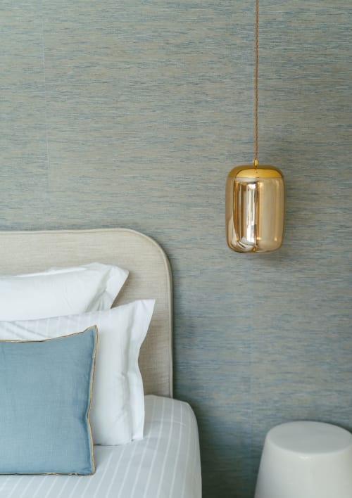 Chambre de la Villa Rhéia avec tête de lit beige, draps blancs, coussin bleu, papier peint bleu-vert texturé et lampe suspendue dorée