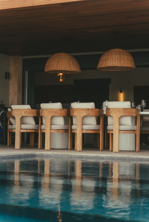 Espace de restauration en bord de piscine à la Villa Théia avec chaises en bois et luminaires suspendus tressés surplombant une eau turquoise