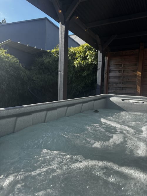 Bain à remous extérieur du gîte d'Armor avec eau bouillonnante, pare-vue en bambou, pergola sombre et porte en bois
