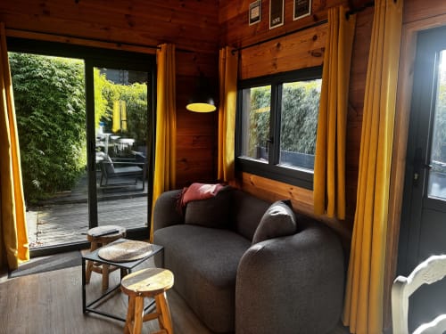 Salon cosy des Gîtes d'Armor avec fauteuil gris, touches de bois, rideaux jaunes et terrasse avec vue sur le jardin