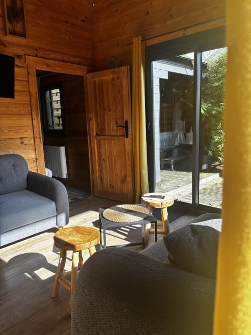 Séjour lumineux avec poutres apparentes, fauteuils gris, tabourets en bois et portes vitrées ouvrant sur la terrasse du jardin aux Gîtes