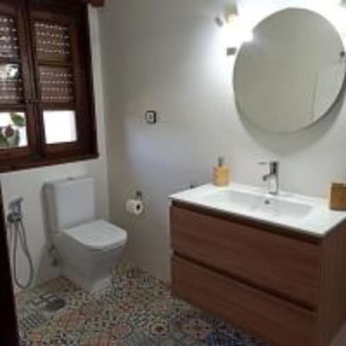 Baño moderno en la Pension Castio de Santillana del Mar con mueble de madera, espejo redondo, inodoro y piso de azulejos