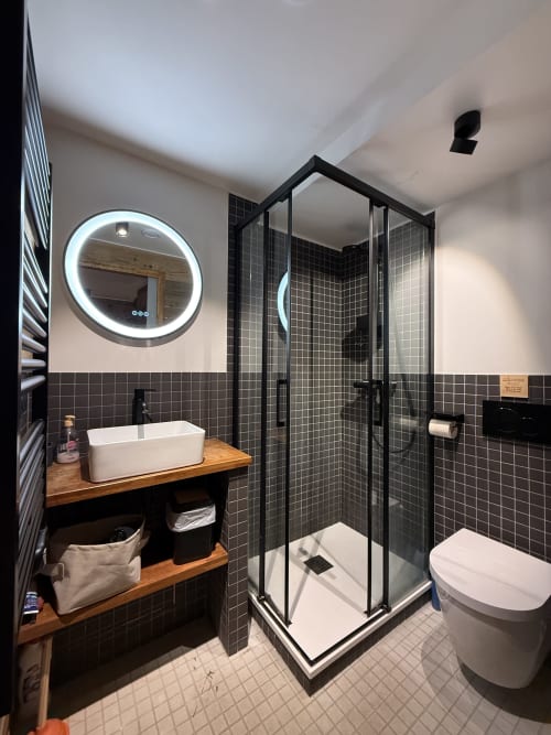 Salle de bain moderne dans un appartement de La Plagne-Tarentaise avec douche carrelée noire, miroir LED, vasque en bois et toilettes