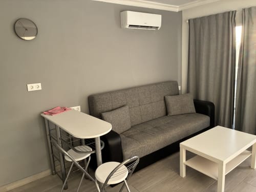 Moderna sala de estar en apartamento ALOE Suite con sofá gris, mesas blancas, cocina americana, aire acondicionado y vista a la piscina