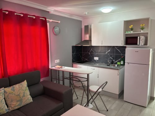 Moderna cocineta con gabinetes rosas, backsplash de mármol negro, mesa de comedor, sofá oscuro y cortinas rojas