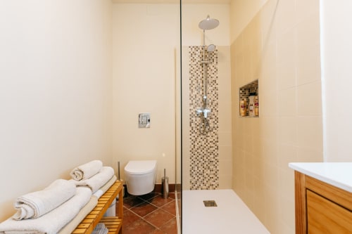 Baño moderno con ducha abierta, pared de azulejos mosaico, inodoro blanco, toallero calefactado y alcachofa de lluvia en La