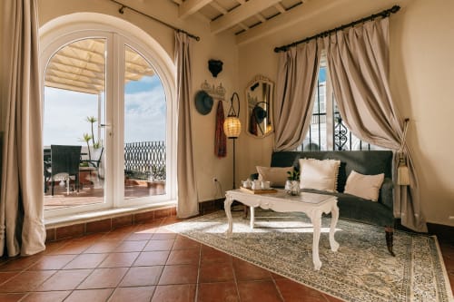 Elegante salón de apartamento estudio con sofá verde, puertas de terraza arqueada con vistas a Medina Sidonia, cortinas color crema y