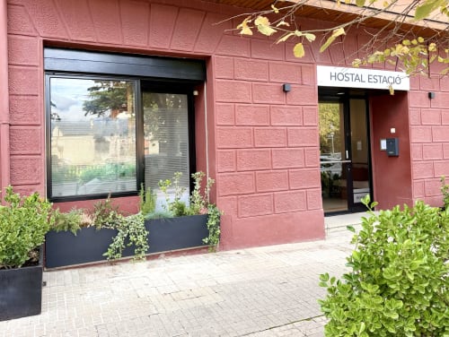 Entrada del Hostal Estació en Puigcerdà con fachada de ladrillo rosa, ventana oscura y macetas con vegetación