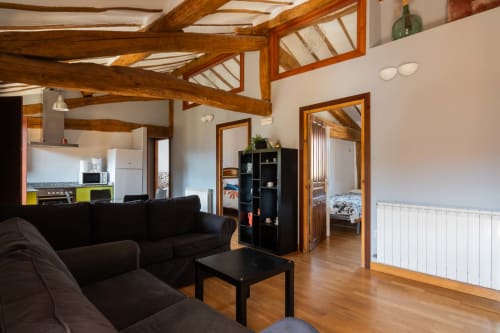 Spacious open-plan living area in La Viña 2 rural apartment with exposed wooden beams, black sofa, kitchen, and bedroom