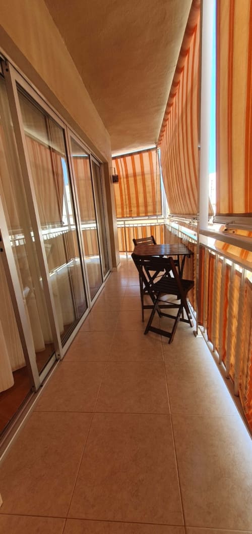 Balcón cubierto angosto con baldosas de terracota, mesita y sillas, toldos rayados naranjas, apartamento Casa Juana en El