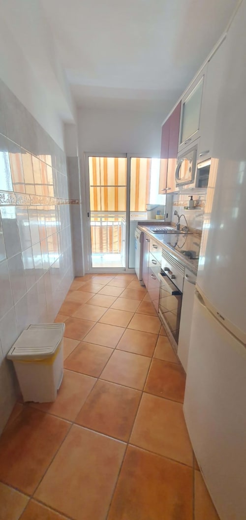 Cocina de galera luminosa en apartamento de El Médano con azulejos de terracota, electrodomésticos blancos y puerta de balcón acristalada con vistas al océano