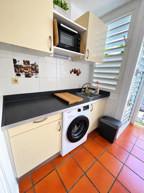 Petite cuisine de Kfé Chocolat avec meubles crème, plan de travail noir, micro-ondes, lave-linge et carrelage en terre cuite.