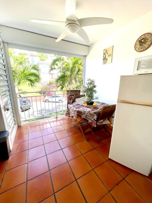 Cuisine lumineuse avec carrelage en terre cuite, ventilateur blanc au plafond, petite table à manger, plante en pot, réfrigérateur et balcon