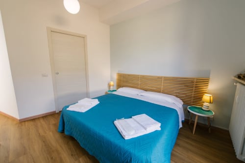 Camera da letto luminosa dell'appartamento di Verona con coperta blu, testata in legno, comodini e porta bianca.
