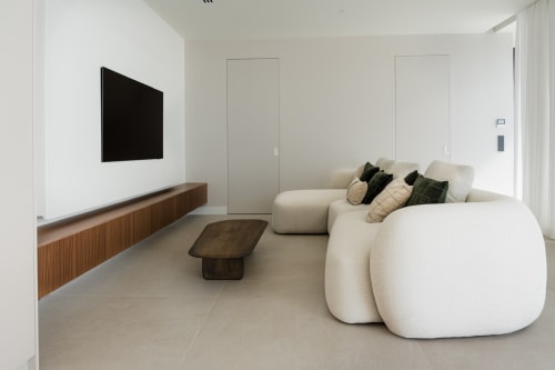 Sala moderna con sofá curvo color crema, TV montado en pared, consola de madera y cojines decorativos en villa V&C Alisio