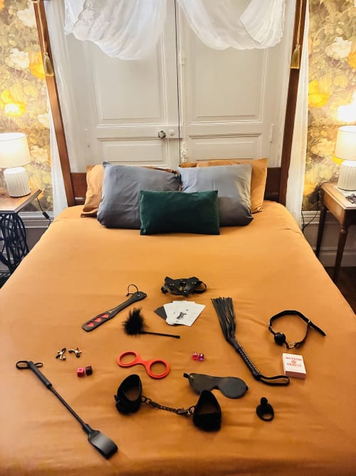 Accessoires intimes pour adultes exposés sur literie orange dans la suite Romance chez Sir Guillaume aux Chez Mary Studios Gannat