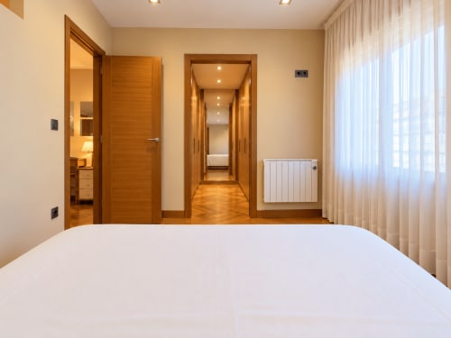 Moderno dormitorio en apartamento de Baiona con puertas de madera, cama blanca y vistas a la ciudad a través de cortinas translúcidas