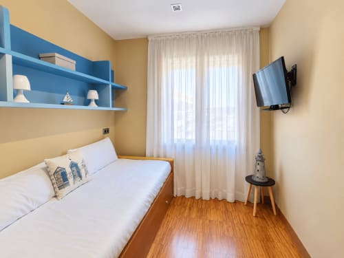 Luminoso dormitorio en Baiona en tonos amarillo y azul, con cama de madera, TV mural, estanterías y vistas a la ciudad