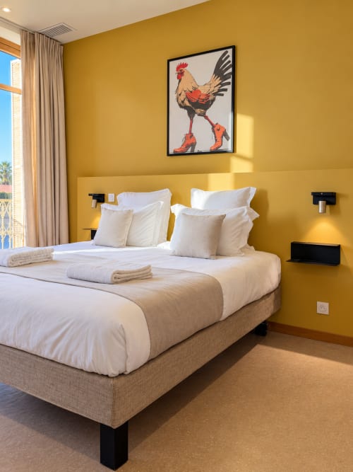 Chambre moderne de l'hôtel Le Palmier Aigues-Mortes avec murs jaune vif, literie blanche et art coq fantaisiste.
