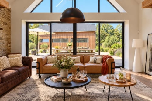 Salon moderne de la villa Domaine Peulven avec canapés en cuir, lampe suspendue noire et grandes baies vitrées donnant sur la campagne