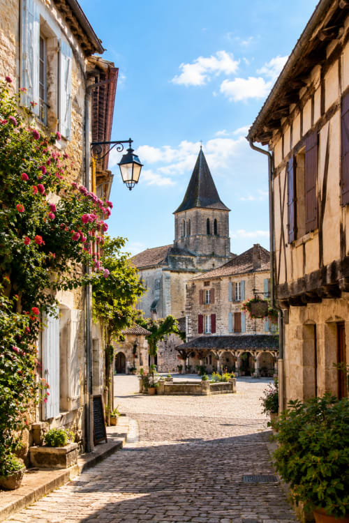 Charmante ruelle pavée d'Eymet, France, bordée de bâtiments couverts de lierre et de fleurs roses, menant à un clocher
