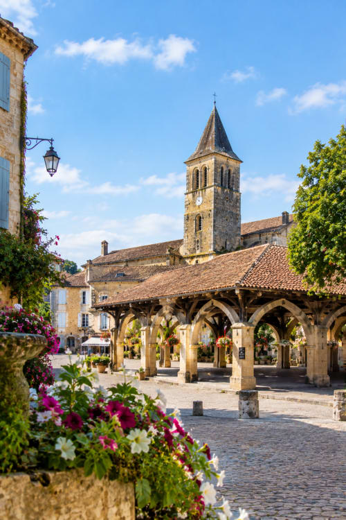 Charmante place de village français à Eymet avec clocher en pierre, marché couvert et fleurs colorées à l'Hôtel les Vieilles
