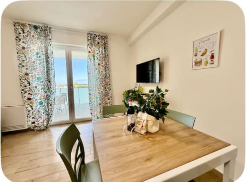 Zona pranzo di moderno appartamento a Bari con vista mare, tavolo in legno, sedie verdi, TV e tende floreali colorate che si aprono su