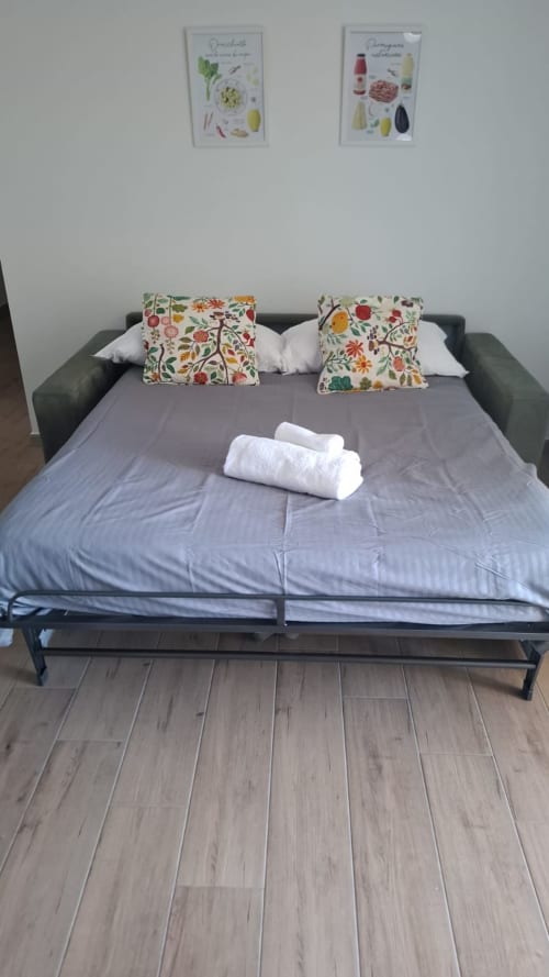 Ampio divano letto presso Newhouse Bari Airport Apartments con cuscini floreali colorati, asciugamani bianchi e biancheria grigia pronta all'uso